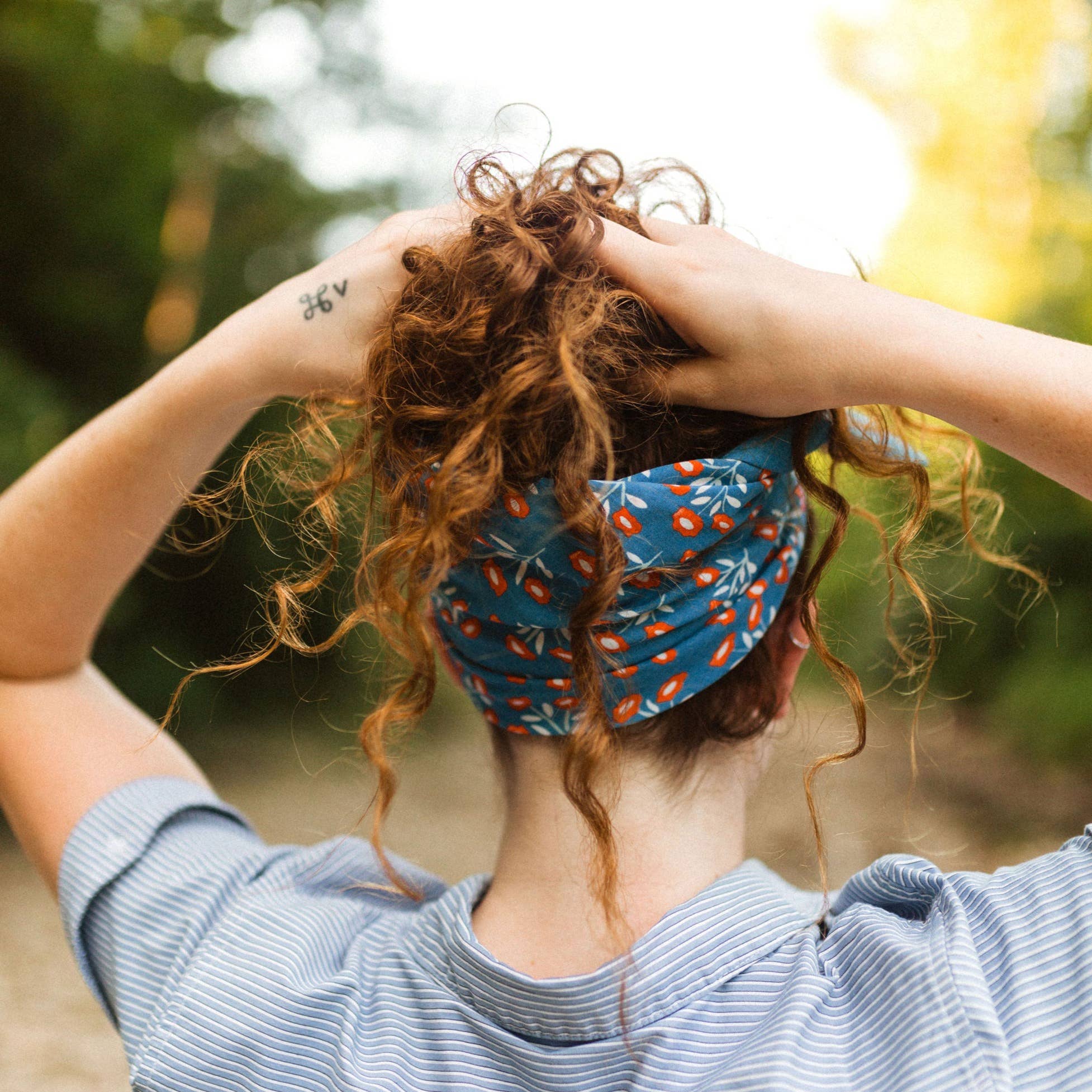 A person with curly hair tied up and covered by the Handker Bandanas No. 075 Juliet Bandana, a hand screen-printed 22x22 inch premium blue cotton accessory with a red and white pattern, stands outdoors in a striped shirt, hands on head.