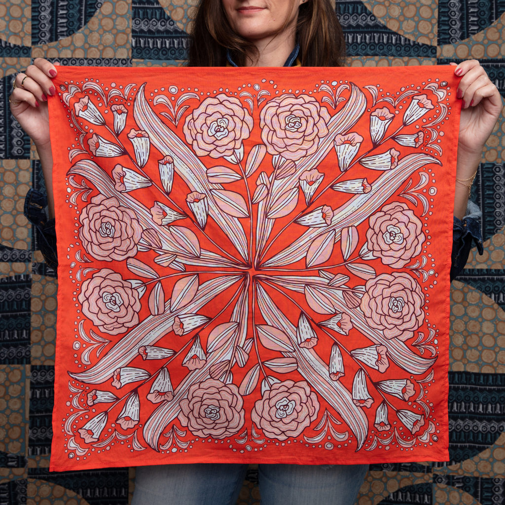 A woman holds the No. 105 Greta Bandana by Handker Bandanas, featuring an intricate floral and leaf pattern with pink flowers and white accents. In the background is a blue and beige patterned textile.