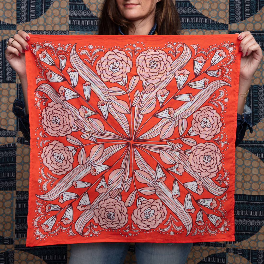 A woman holds the No. 105 Greta Bandana by Handker Bandanas, featuring an intricate floral and leaf pattern with pink flowers and white accents. In the background is a blue and beige patterned textile.