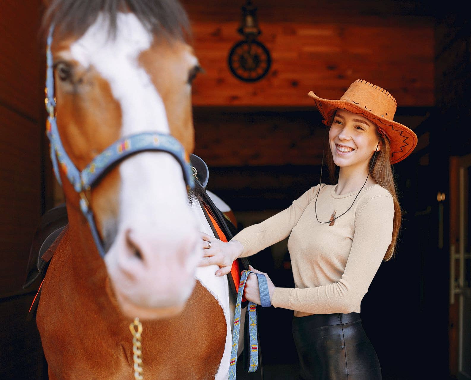 A smiling woman in a cowboy hat stands next to a brown and white horse in a stable, her jacket featuring the Chestnut Horse Head - Facing Left Iron on Patch by Wholesale Applique, highlighted by warm light in the wooden interior.