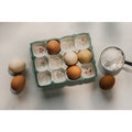 The Danica Heirloom Camellia Egg Holder, adorned with floral patterns, holds five eggs. Three more eggs and a metal measuring cup of flour rest nearby on a white textured surface.