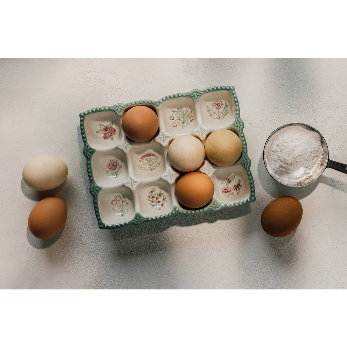 The Danica Heirloom Camellia Egg Holder, adorned with floral patterns, holds five eggs. Three more eggs and a metal measuring cup of flour rest nearby on a white textured surface.
