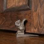 The 3.5" METAL SQUIRREL DOOR STOP by Ragon House, designed as a squirrel holding a nut, rests at the base of a wooden door on a hardwood floor.