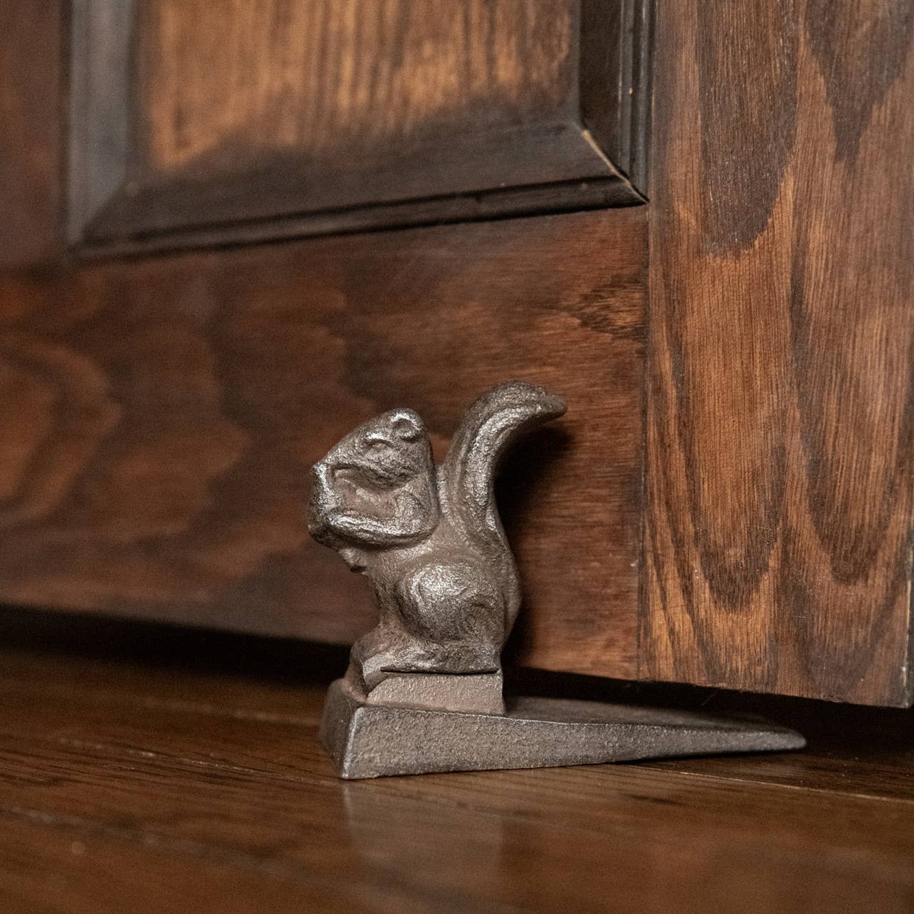The 3.5" METAL SQUIRREL DOOR STOP by Ragon House, designed as a squirrel holding a nut, rests at the base of a wooden door on a hardwood floor.