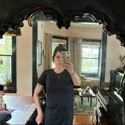 A woman with dark hair and glasses takes a mirror selfie in a cozy, sunlit living room. She’s wearing the Basic Black Crop from Thyne Goods and holding her phone with a floral case.