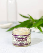 An open jar of Roots And Leaves Magnesium Body Butter rests on a light surface, with a blurred green plant and glass bottle behind it. The label showcases fast relief, magnesium absorption, and nourishing shea, cocoa, and mango butters.
