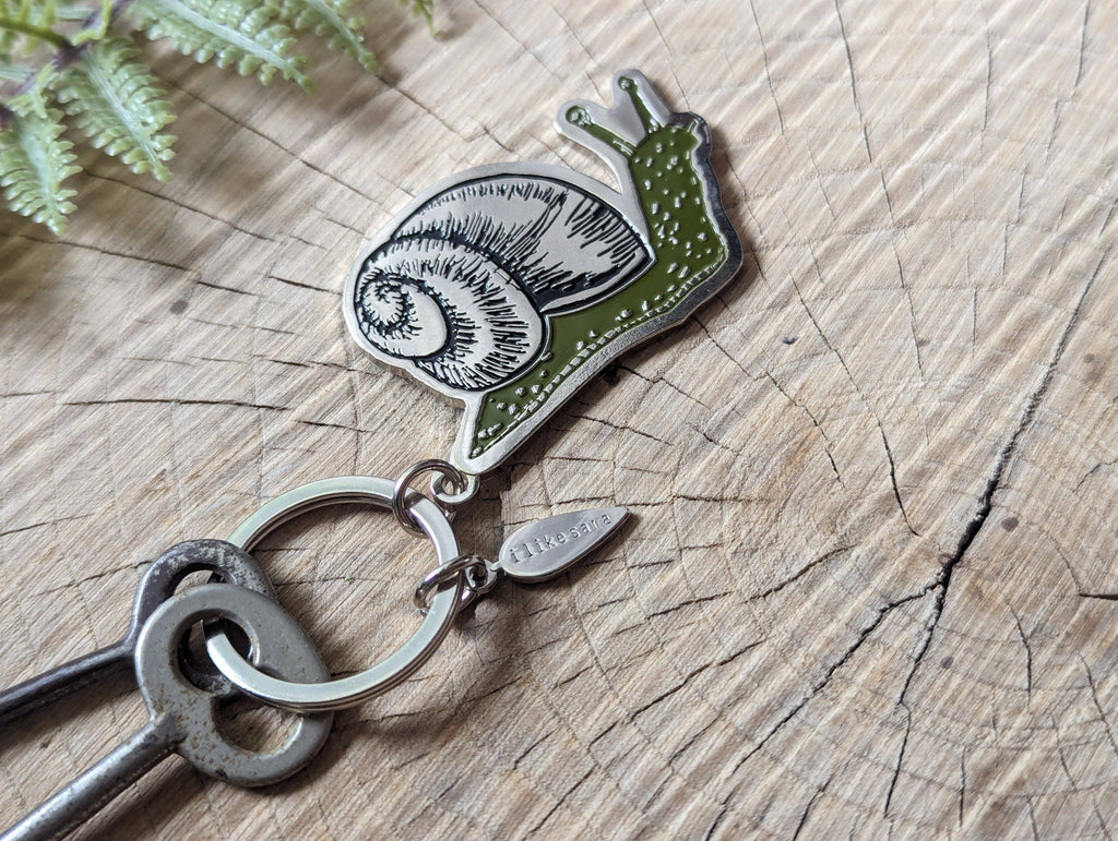 The Metal enamel keychain / purse charm | snail by ilikesara | art + goods features a detailed green snail charm and a silver-plated tag engraved with hkgifts, displayed on weathered wood with fern leaves nearby.
