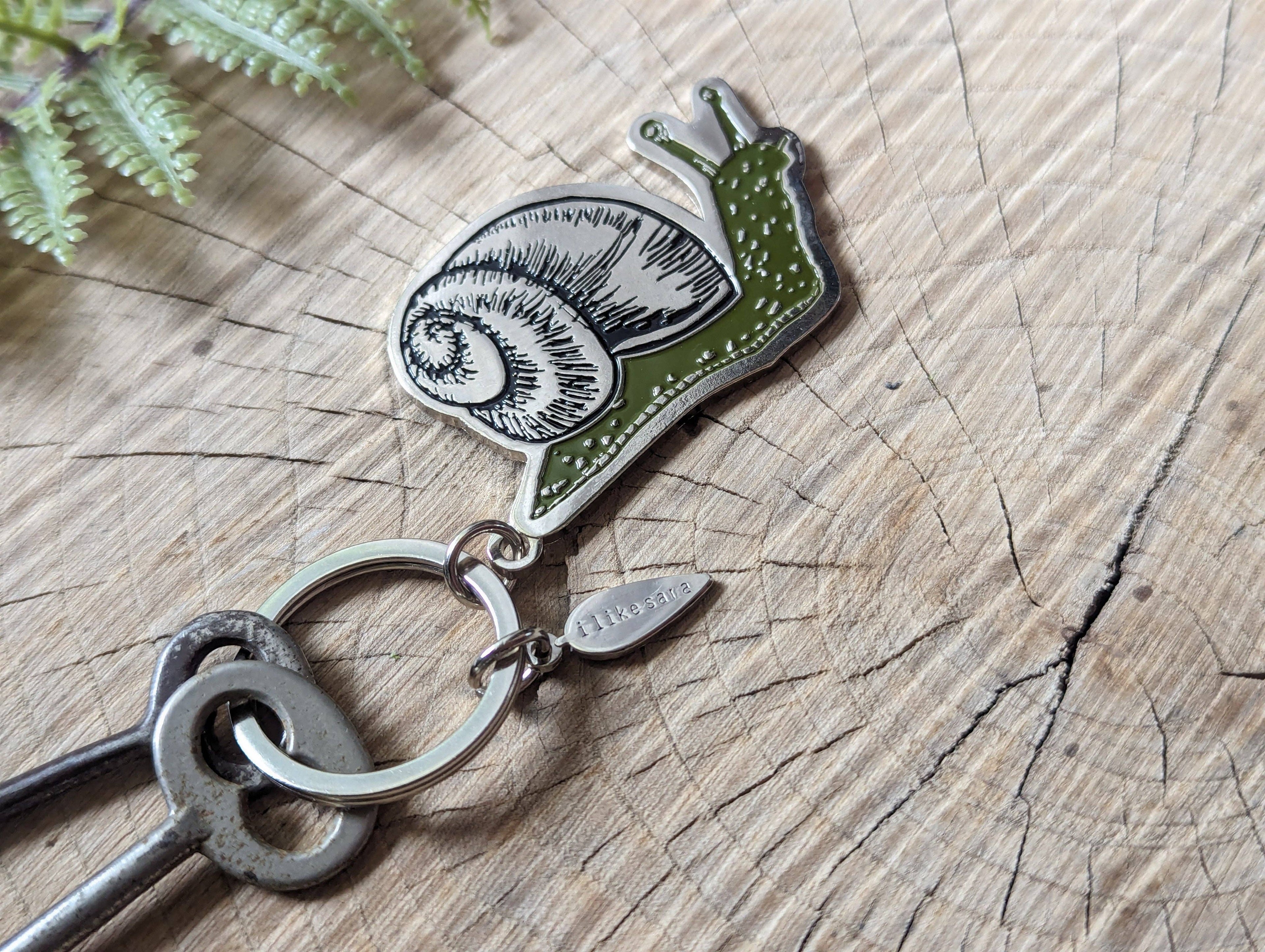 The Metal enamel keychain / purse charm | snail by ilikesara | art + goods features a detailed green snail charm and a silver-plated tag engraved with hkgifts, displayed on weathered wood with fern leaves nearby.