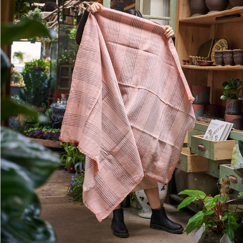 A person stands indoors holding up the Variously Silica Rose Throw in shades of salmon gray stripes, concealing their face and body, surrounded by plants and shelves filled with pots and gardening supplies.