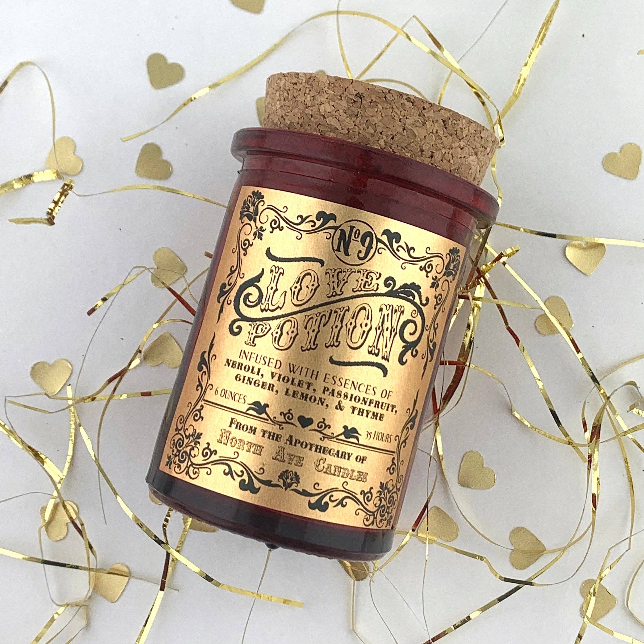 The Apothecary / Love Potion / Valentine Candle by North Ave Candles features a red glass jar with a vintage label, surrounded by gold heart confetti and ribbon, and offers a complex blend of ginger, lemon, passionfruit, and thyme essences.