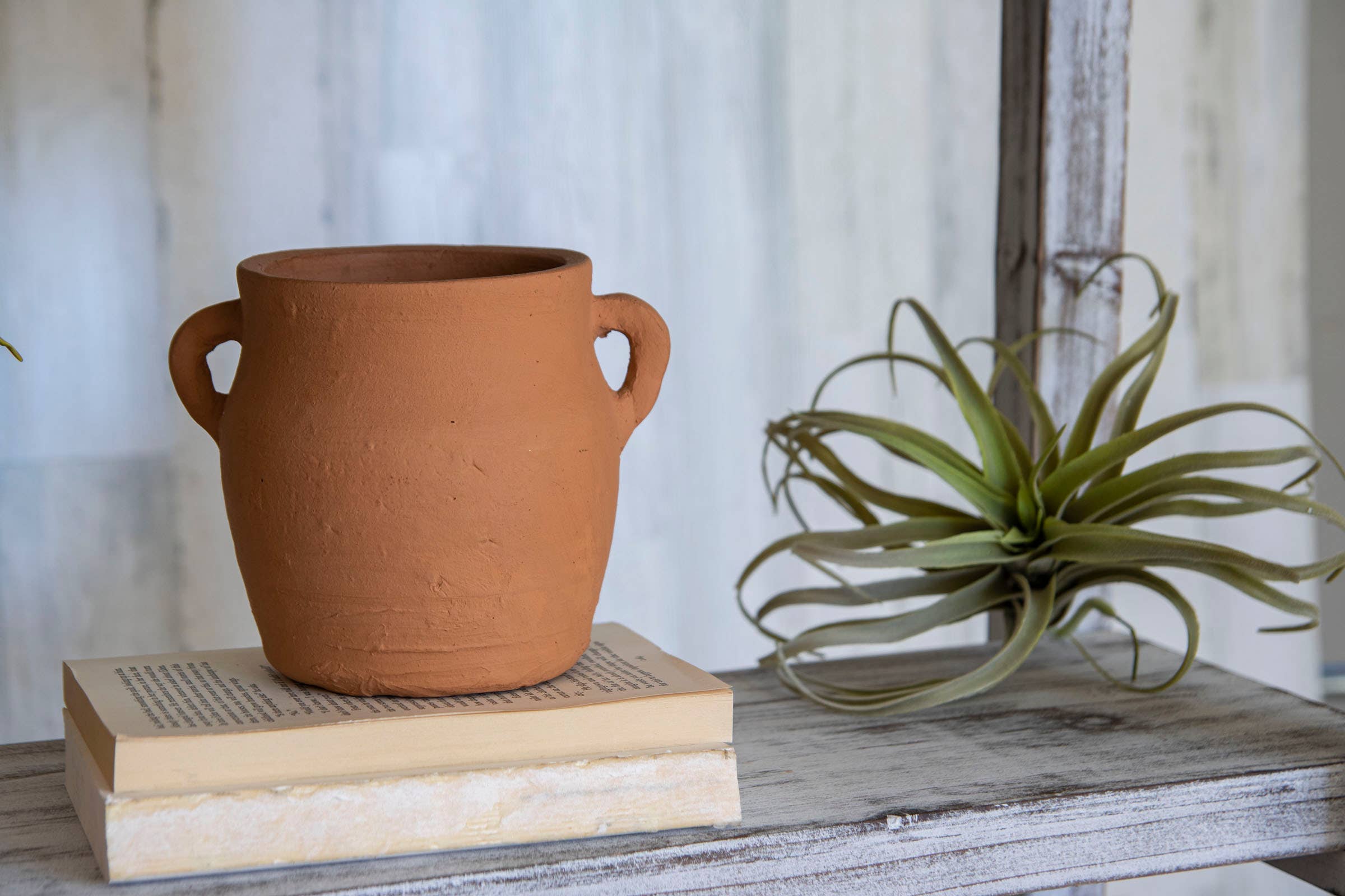 The Foreside Home & Garden Natural Hand Thrown Terracotta Vase with Handles rests atop a closed book beside a leafy air plant on a rustic wooden shelf, set against a light, textured background.