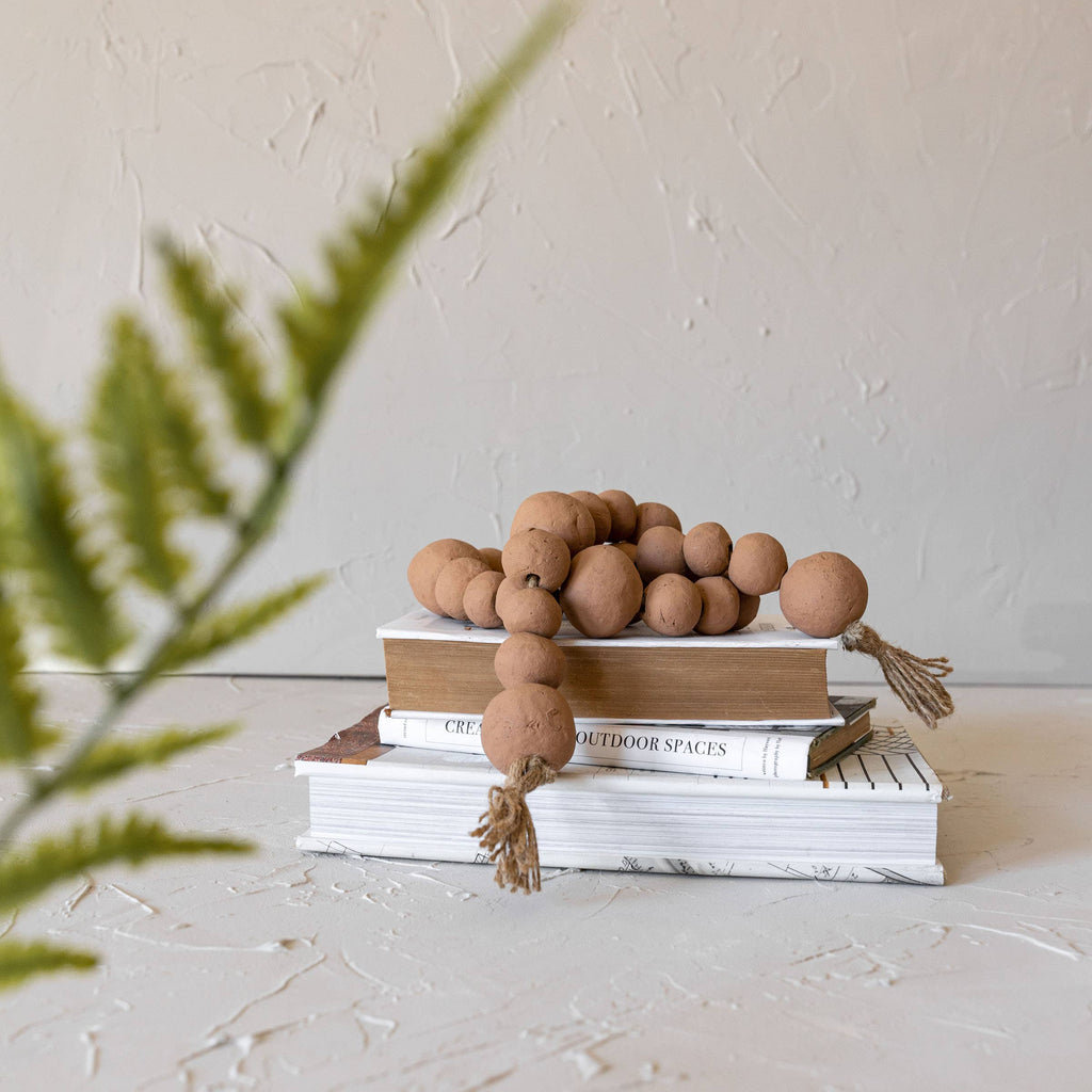 A stack of books topped with the Clay Garland Terracotta + Jute by Foreside Home & Garden, artisan-made in India, rests on a light textured surface with a blurred green fern frond in the foreground.