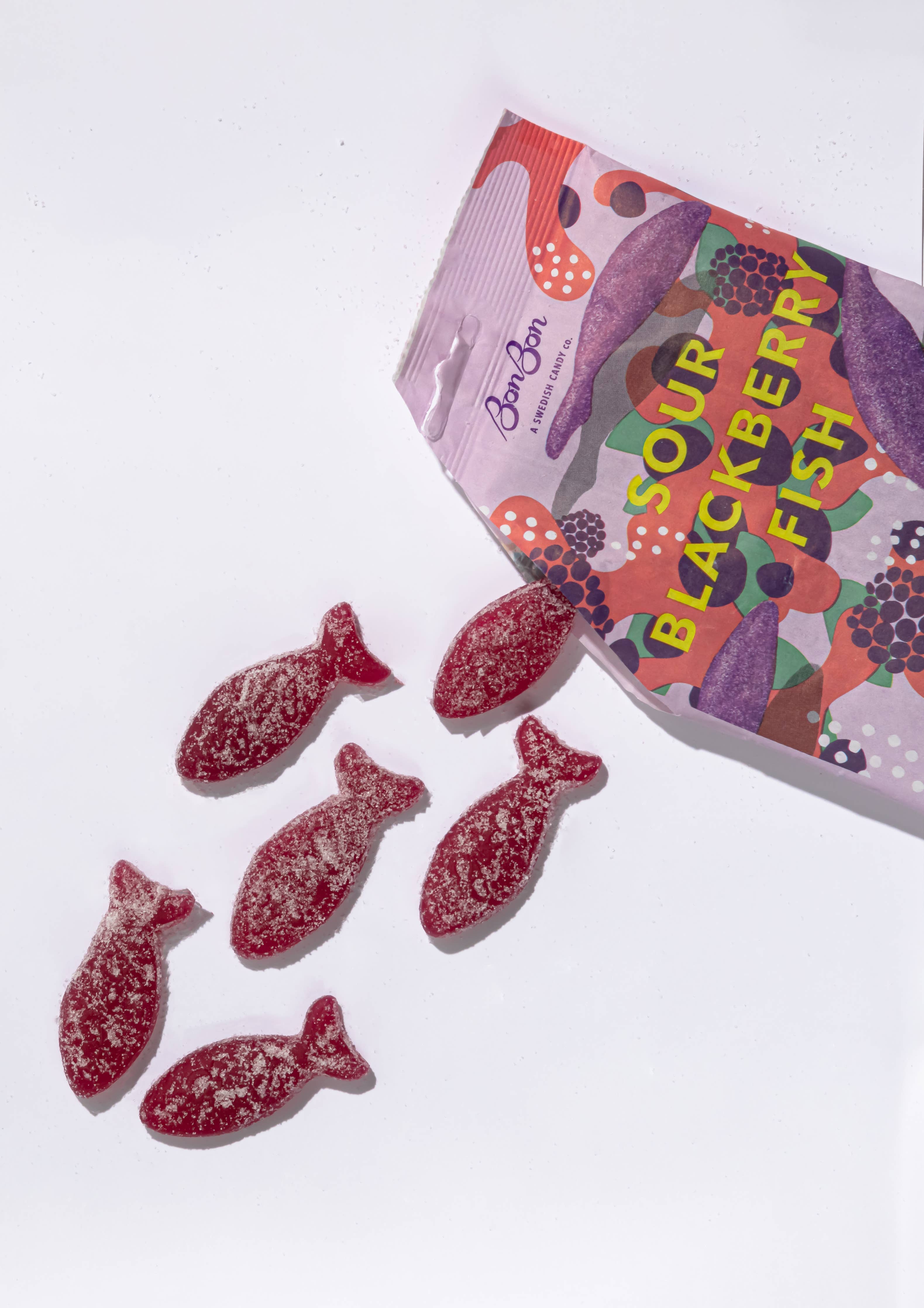 An open bag of BonBon - A Swedish Candy Co Sour Blackberry Fish - Swedish Candy 5.2oz (150g) spills six sugar-coated, vegan, fish-shaped candies onto a white background—offering a fruity twist on classic Swedish Fish.