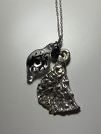 The Broken Bird necklace by Thyne Goods features two pendants: a clear glass art charm with a black eye and leaf design, and an abstract sculpted recycled sterling silver piece. Collaboration by Ash Espinoza x Thyne Studio.
