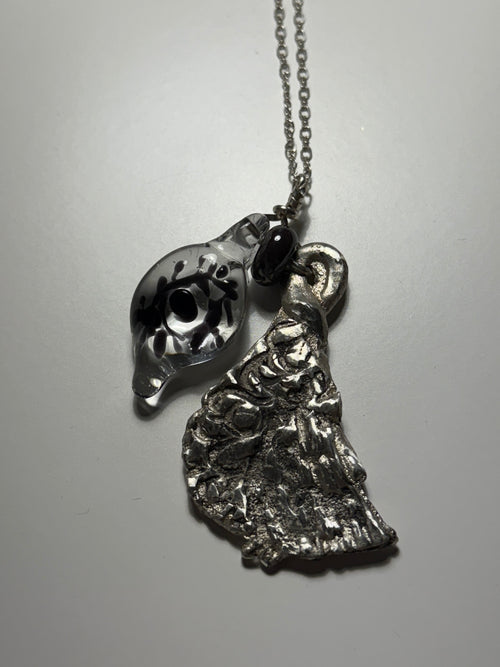 The Broken Bird necklace by Thyne Goods features two pendants: a clear glass art charm with a black eye and leaf design, and an abstract sculpted recycled sterling silver piece. Collaboration by Ash Espinoza x Thyne Studio.