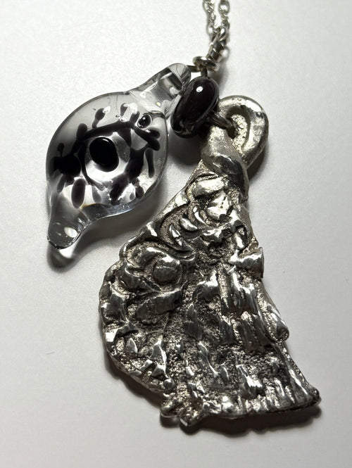 The Broken Bird necklace by Thyne Goods features glass art—a clear pendant with a black dot and abstract design—and a textured, recycled sterling silver face pendant. Collaboration: Ash Espinoza x Thyne Studio.