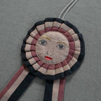 The Skippy Cotton Prize Ribbon - Cotton filled Ornament features a hand-embroidered human face with blue eyes, red lips, and yellow eyebrows on a gray textured backdrop—a unique piece for handcrafted decor.