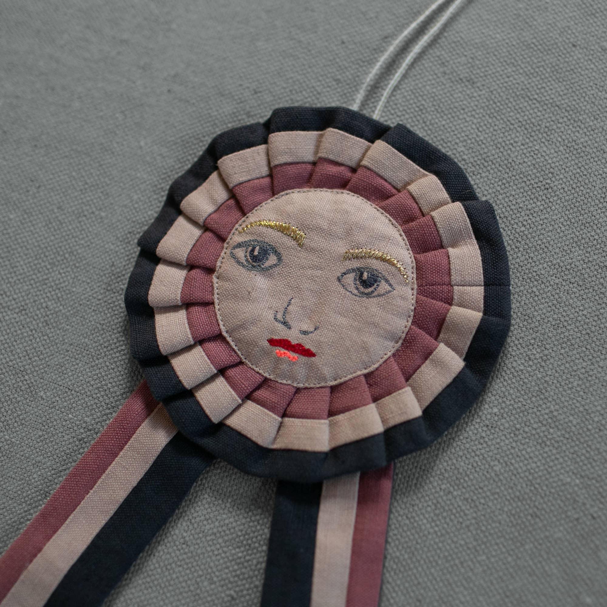 The Skippy Cotton Prize Ribbon - Cotton filled Ornament features a hand-embroidered human face with blue eyes, red lips, and yellow eyebrows on a gray textured backdrop—a unique piece for handcrafted decor.