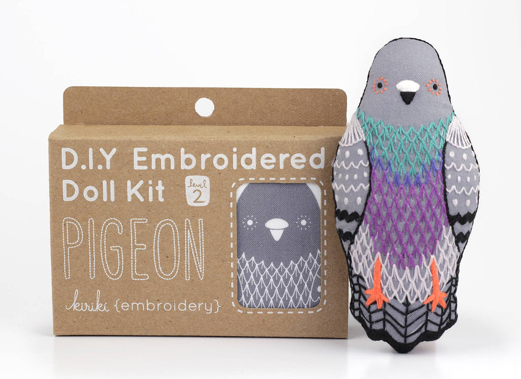 A Pigeon - Embroidery Kit in a cardboard box, standing next to a finished pigeon doll with grey, purple, and green screen-printed feathers.
