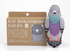 A Pigeon - Embroidery Kit in a cardboard box, standing next to a finished pigeon doll with grey, purple, and green screen-printed feathers.