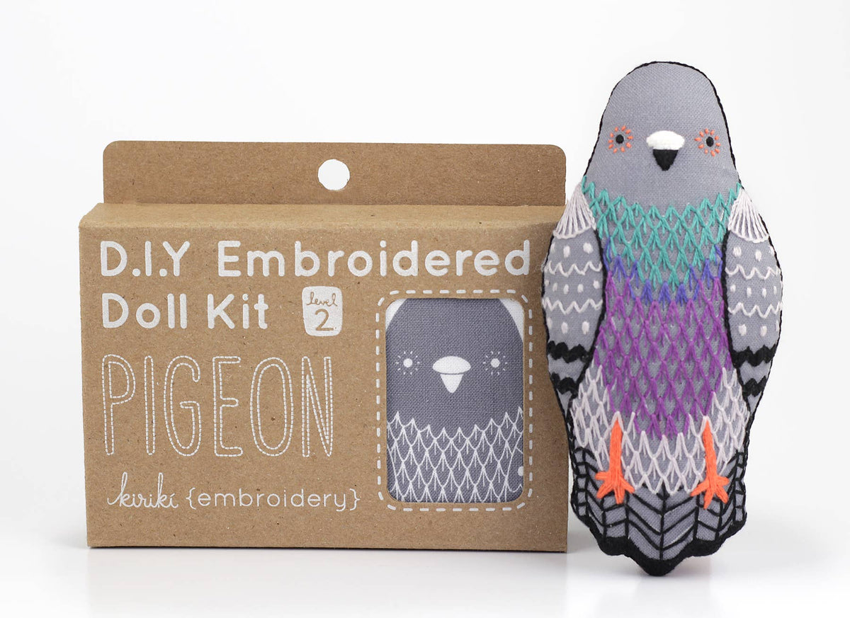 A Pigeon - Embroidery Kit in a cardboard box, standing next to a finished pigeon doll with grey, purple, and green screen-printed feathers.