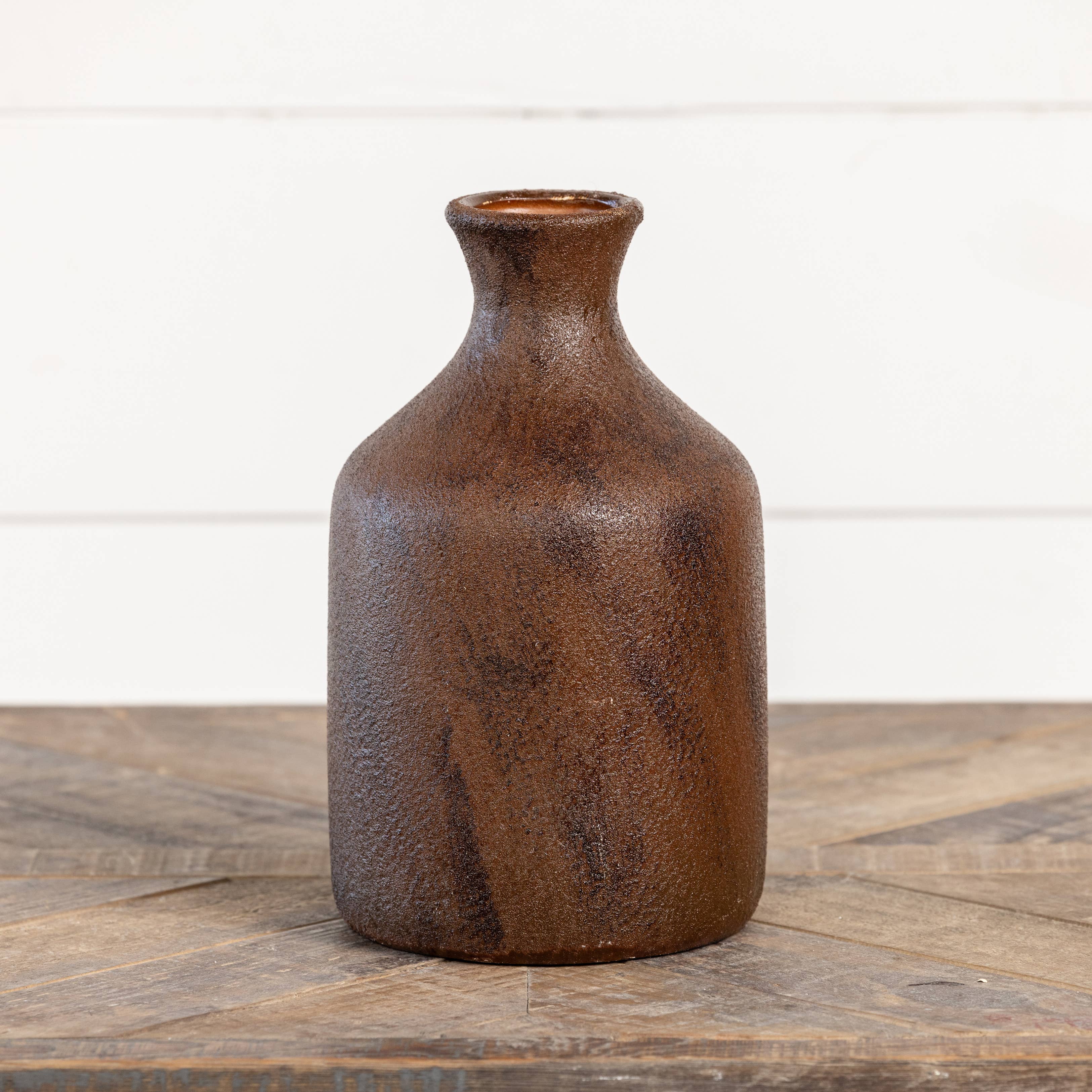 The Ragon House 9.75" Brown & Rust Tone Vase, featuring a textured brown finish and slender neck, sits on a wooden surface against a white background.