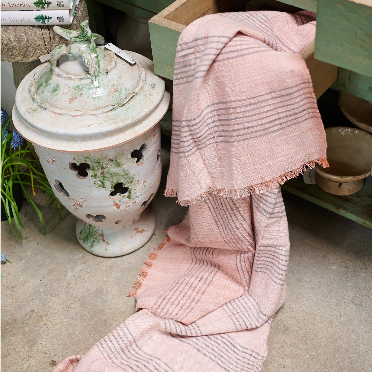 A Silica Rose Throw in shades of salmon and gray stripes by Variously, featuring fringed edges, is draped over the edge of a wooden shelf above a ceramic garden stool with cutout detailing, next to pottery and a concrete floor.