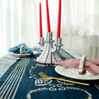 A dining table set with the Ichcha Deepest Navy & White Table Linens Cotton Table Runner - Danya, two plates with pink and blue napkins in white rings, gold forks, a white candelabra holding three red candles, as soft light filters through curtains.