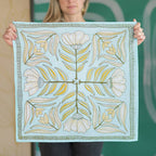 A woman holds up the Handker Bandanas No. 012 Eliza Bandana, a light blue premium cotton accessory featuring a white, green, and yellow symmetrical floral design with four large flowers and radiating leaves for an elegant touch.