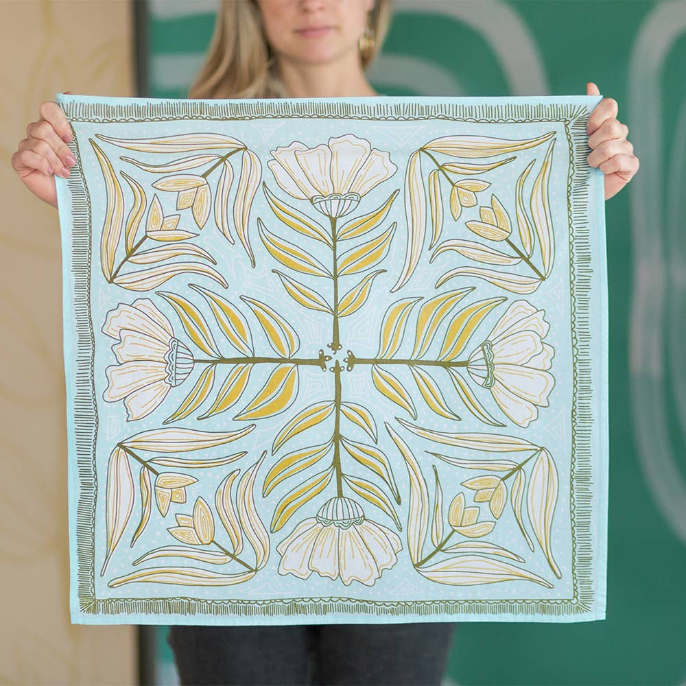 A woman holds up the Handker Bandanas No. 012 Eliza Bandana, a light blue premium cotton accessory featuring a white, green, and yellow symmetrical floral design with four large flowers and radiating leaves for an elegant touch.