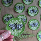 A hand holds the ilikesara | art + goods Iron-on patch | polyphemus moth with lichen - green, featuring a black and white moth on fern-green. Identical patches are displayed on wood alongside real fern leaves for insect-themed flair.