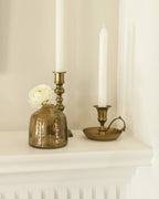 A blush Bella Handblown Hammered Glass Bud Vase by Creative Women with a small white flower sits on a mantel beside two brass candle holders with white taper candles against a white wall.