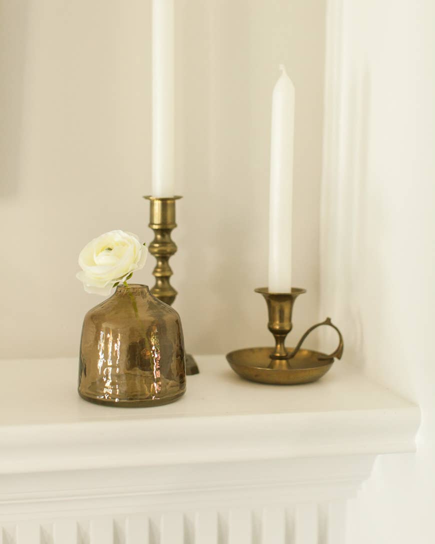 A blush Bella Handblown Hammered Glass Bud Vase by Creative Women with a small white flower sits on a mantel beside two brass candle holders with white taper candles against a white wall.