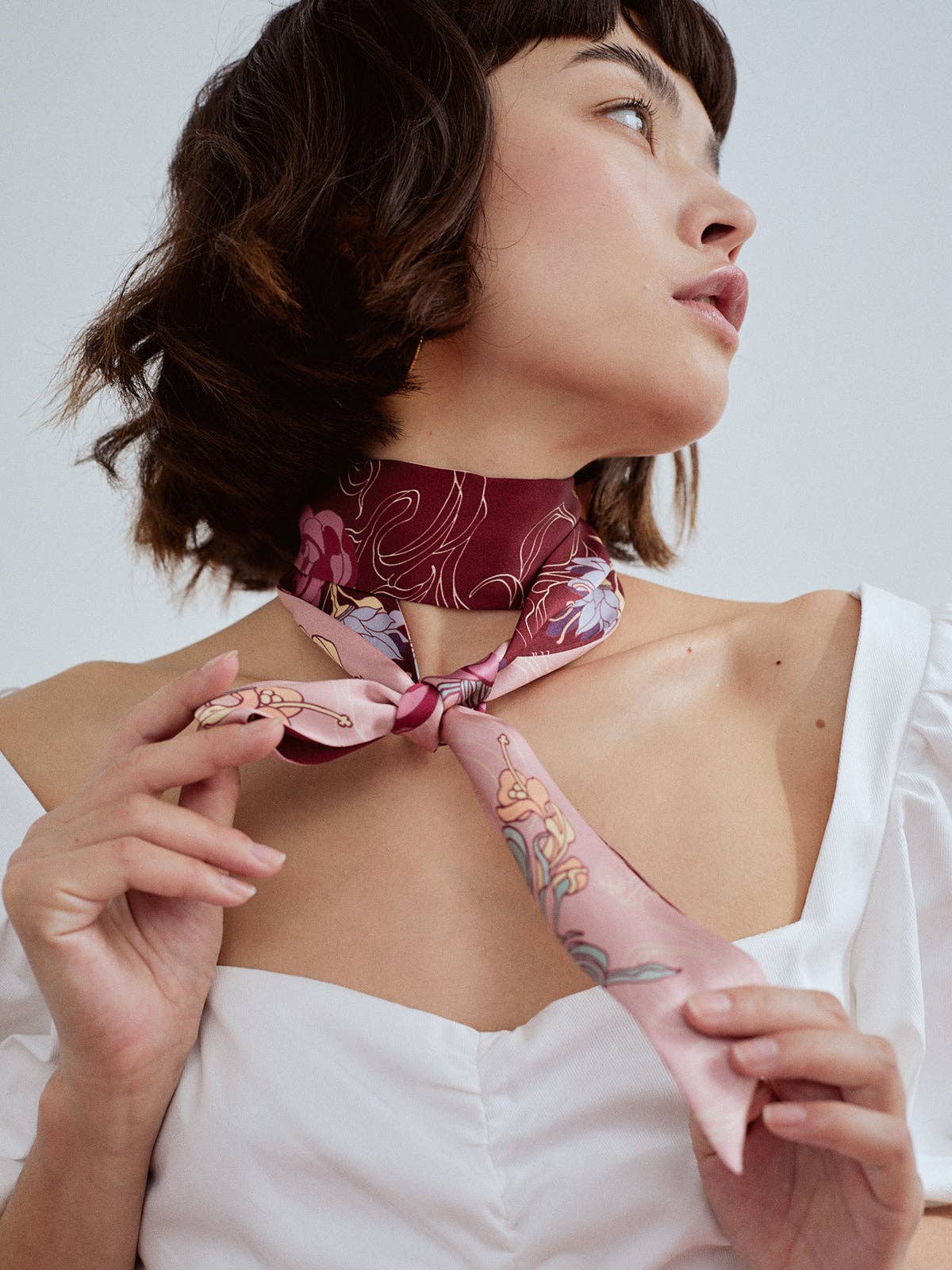 A woman in a white off-shoulder top adjusts the Lost Pattern NYC Silk Skinny Scarf - Ruby Red around her neck. This chic mulberry silk scarf can also be worn as a stylish hair accessory or headband for added versatility.
