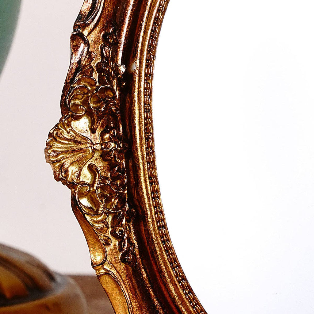Close-up of Vagabond Vintage's Vintage Style Ornate Oval Table Mirror (Medium) featuring an intricate gold floral frame against a white background, with a hint of green and gold in the corner—ideal for vintage vanity settings.