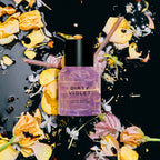 A 15ML bottle of Heretic Parfum's DIRTY VIOLET rests on a glossy black surface, surrounded by scattered yellow, pink, and white petals, creating an artistic scene that hints at patchouli and violet leaf notes.