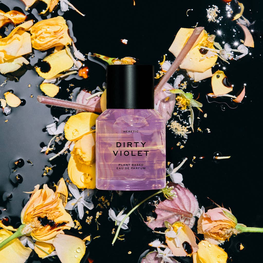 A 15ML bottle of Heretic Parfum's DIRTY VIOLET rests on a glossy black surface, surrounded by scattered yellow, pink, and white petals, creating an artistic scene that hints at patchouli and violet leaf notes.