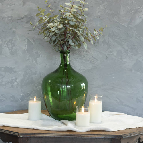 The Ragon House 16" GREEN GLASS BOTTLE, filled with eucalyptus, creates a stunning centerpiece on a white-clothed wood table, surrounded by three lit white candles in glass holders against a textured gray wall.