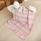 The Silica Rose Throw in Shades of Salmon Gray Stripes Pattern by Variously is draped over a white oval coffee table on a light wood floor, next to a brown sofa and a decorative vase.