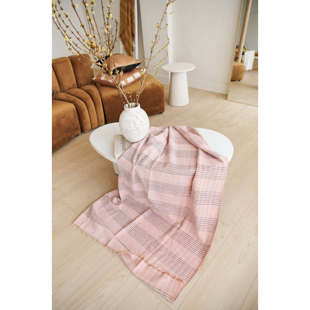 The Variously Silica Rose Throw in shades of salmon with a gray stripes pattern is draped over a white coffee table, next to a white vase with branches, in a modern living room featuring light wood floors and a brown couch.