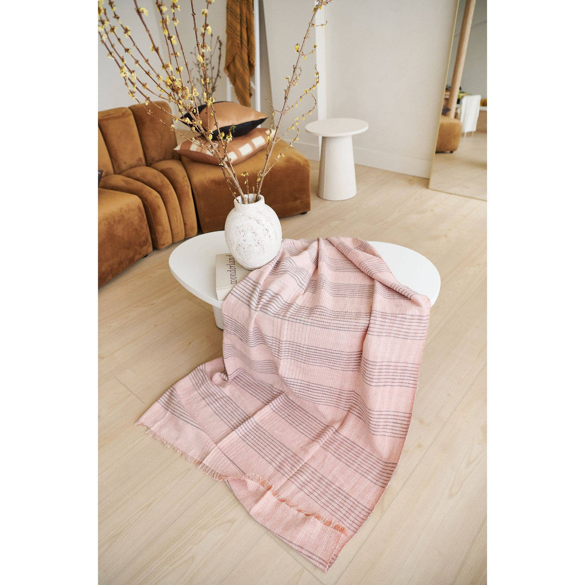 The Variously Silica Rose Throw in shades of salmon with a gray stripes pattern is draped over a white coffee table, next to a white vase with branches, in a modern living room featuring light wood floors and a brown couch.