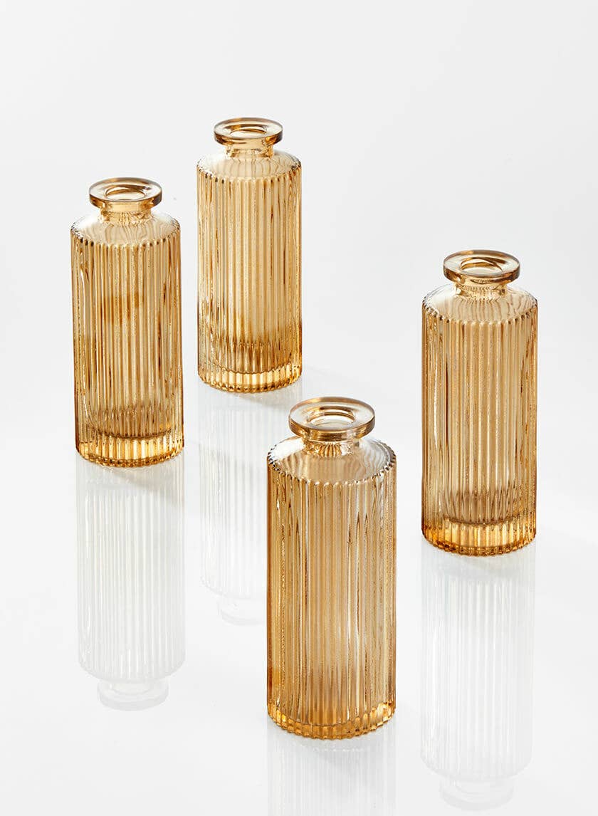 Four Jamali Garden Amber Pleated Glass Bottle Bud Vases with narrow necks are arranged on a reflective white surface, casting gentle shadows and reflections.