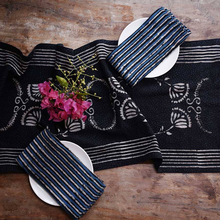Two white plates with folded striped blue cloth napkins sit on a wooden table featuring Ichcha’s Deepest Navy & White Table Linens Cotton Table Runner – Danya, accented by a small bouquet of pink flowers near the top left plate.