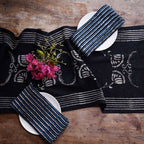 Two white plates with folded striped blue cloth napkins sit on a wooden table featuring Ichcha’s Deepest Navy & White Table Linens Cotton Table Runner – Danya, accented by a small bouquet of pink flowers near the top left plate.