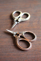 Two pairs of NNK Press Mini Embroidery Scissors: Antique Gold, with stainless steel blades and ornate handles, rest on a wooden surface—ideal compact sewing snips for your travel notions kit.