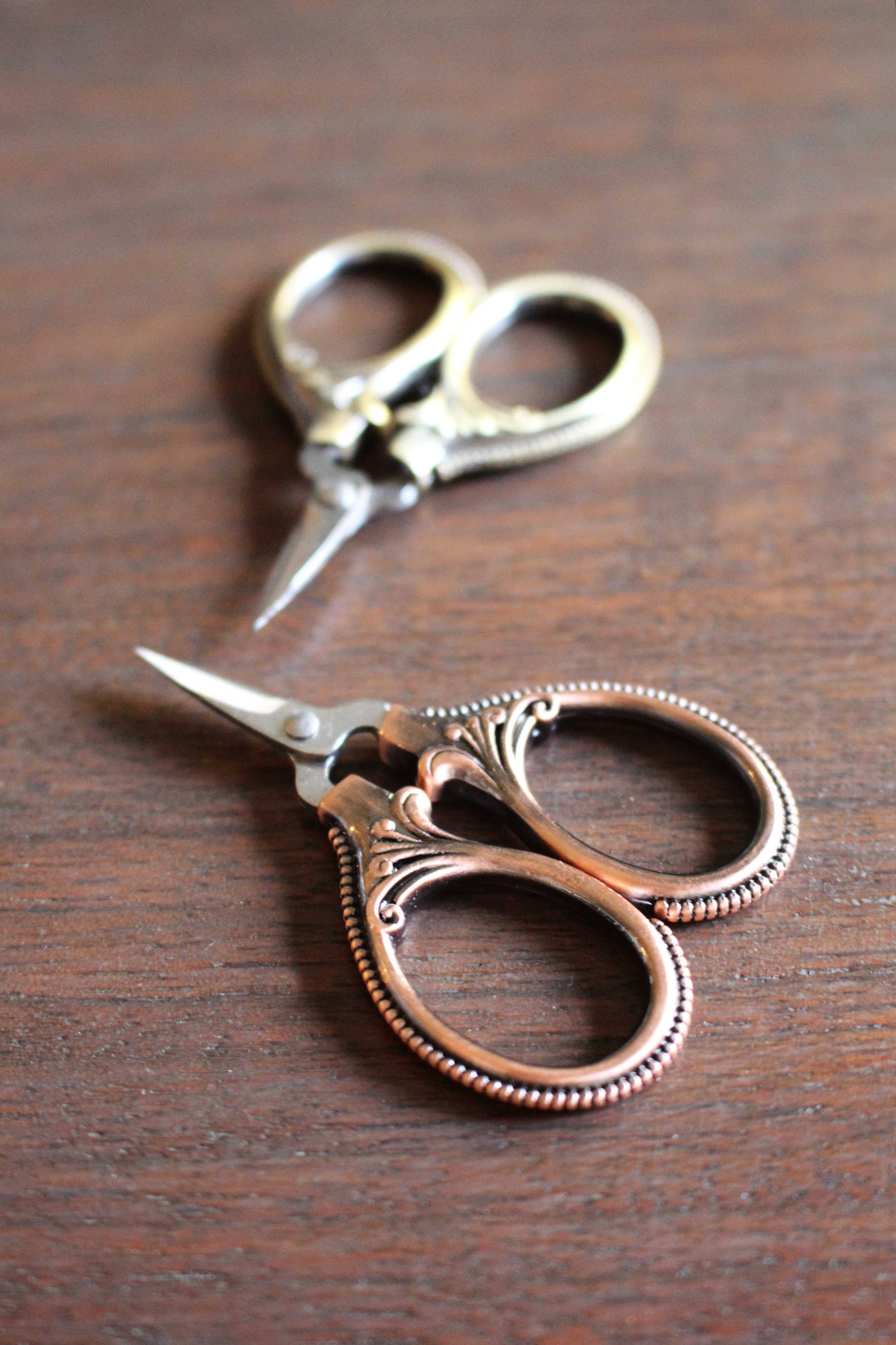Two pairs of NNK Press Mini Embroidery Scissors: Antique Gold, with stainless steel blades and ornate handles, rest on a wooden surface—ideal compact sewing snips for your travel notions kit.