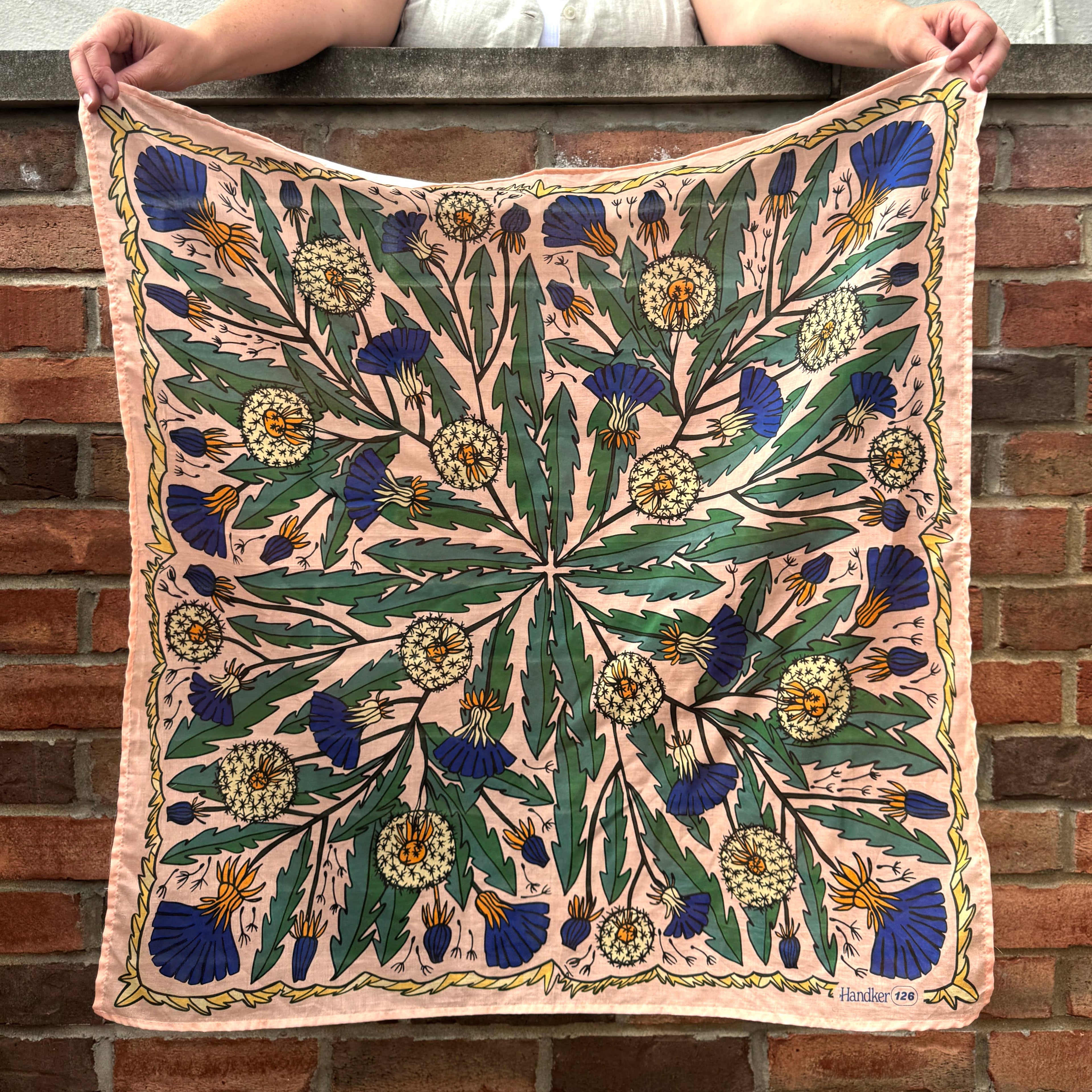 A person holds up the Handker Bandanas 34" Dandy Wild Rag Bandana, featuring a premium cotton square with green leaves, yellow and white flowers, and blue thistles on pale pink, standing before a red brick wall.