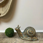 The Vagabond Vintage CAST IRON SNAIL STATUE: Gold rests on a rustic wood surface next to a green moss ball, adding charm to your home decor; a light wall and wooden frame appear in the background.