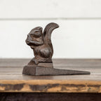 The 3.5" METAL SQUIRREL DOOR STOP by Ragon House sits upright on its wedge base, displayed on a wooden surface against a light background.