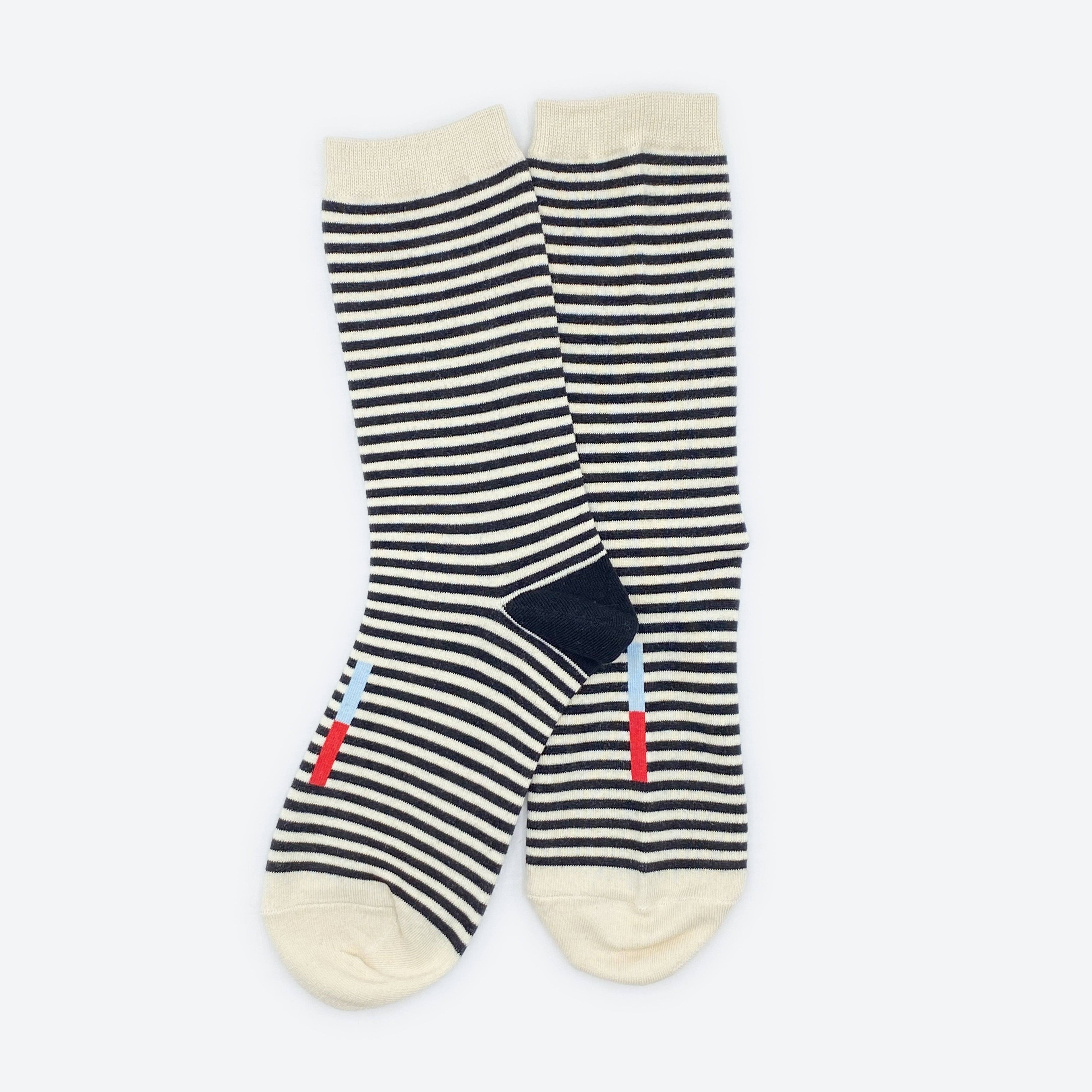 The Cole Black and White Striped Crew Socks by Hooray Sock Co. are unisex crew socks with cream toes, heels, and cuffs, plus small blue and red stripes near the bottom.